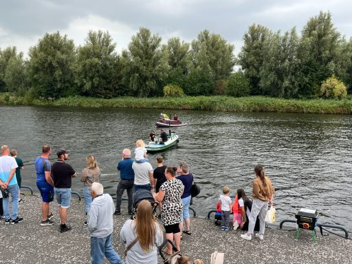 Mensen kijken naar een boot op een rivier, omringd door bomen en een bewolkte lucht.