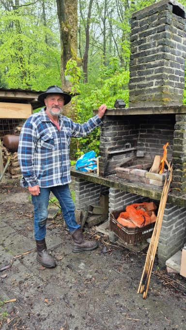Een man met een hoed staat naast een buitenoven in een groene bosomgeving.