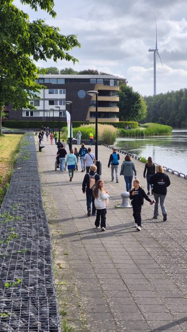 Wandelende groep mensen langs een laan bij een waterweg, met bomen en gebouwen op de achtergrond.