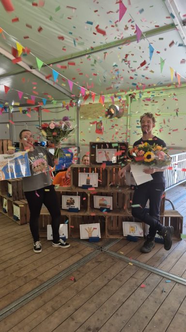 Twee mensen staan tussen versierde dozen met feestdecoratie en een kleurrijke achtergrond.