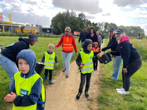 Groep mensen en kinderen in gele hesjes verzamelen afval in een groenzone.