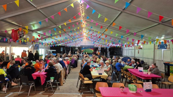 Drukke feesttent met kleurrijke vlaggen en mensen aan lange tafels.