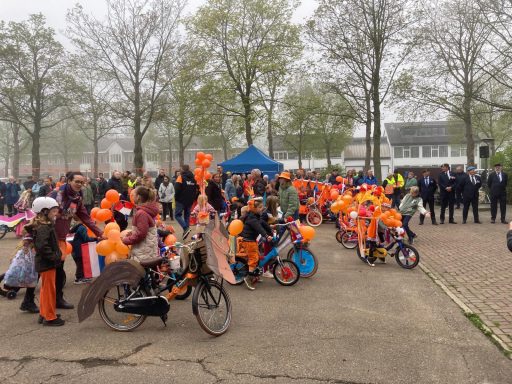 Een groep kinderen in oranje kleding met fietsen en ballonnen, omringd door een menigte.