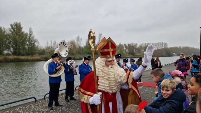 Sinterklaas groet kinderen bij het water, omringd door muzikanten en feestelijke sfeer.