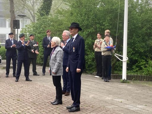 Ceremonie met mensen in formele kleding bij een vlaggenmast, publiek applaudisseert.