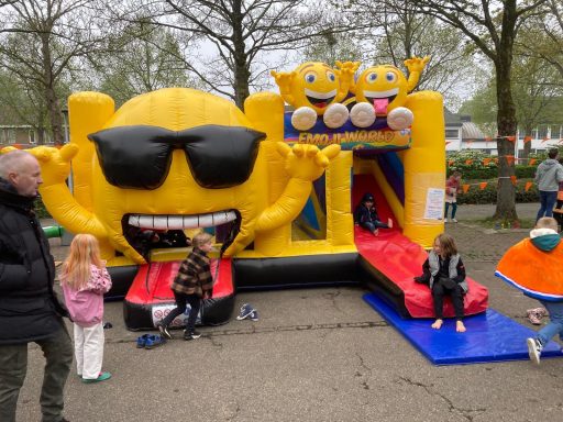 Gele opblaasbare glijbaan met zon en lachende gezichten, kinderen spelen erbij.